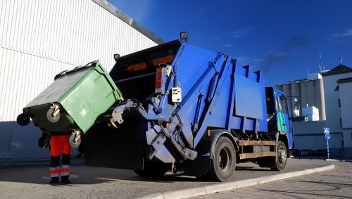 A large blue refuse collection truck with a hydraulic lifting mechanism at the rear is shown in an outdoor urban setting, positioned on a paved street next to a white industrial building. The truck is actively in the process of emptying a green wheeled rubbish bin into the container at the back, with the binâ€™s lid slightly ajar. The bin, made of durable plastic, has a textured surface and grey wheels, and stands on the ground with its handle and wheels visible. The driverâ€™s cabin is on the right side of the vehicle, with reflective safety strips and side mirrors. The scene is illuminated by daylight, with a clear blue sky overhead, and the area appears clean and well-maintained, characteristic of a municipal waste collection service in the vicinity of Deptford or nearby London districts. The overall setting emphasizes professional rubbish removal activities typical of services provided by companies like Office Clearance Deptford, which operate within the London boroughs for waste collection and clearance tasks.