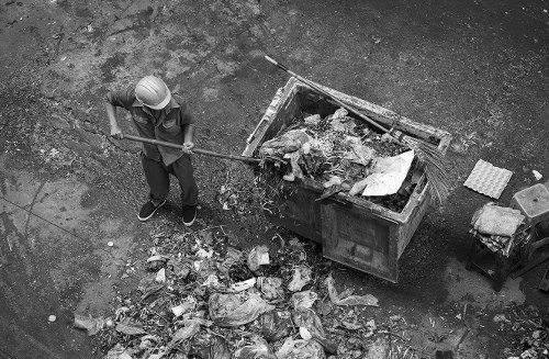 A worker wearing a protective helmet and casual clothing is using a long-handled tool to clear rubbish from a large metal skip container positioned outdoors on a gravel surface. The rubbish inside the skip includes various debris such as paper, plastic, and unidentifiable waste materials. Some waste has spilled onto the ground nearby, forming a small pile of mixed refuse. In the background, there are additional items including a small chair or stool and concrete blocks or paving slabs arranged on the ground. The scene appears to be part of an outdoor rubbish removal operation, likely associated with a professional service such as Office Clearance Deptford, which services the local area around Deptford or SE postcodes, offering waste management and rubbish clearance solutions.