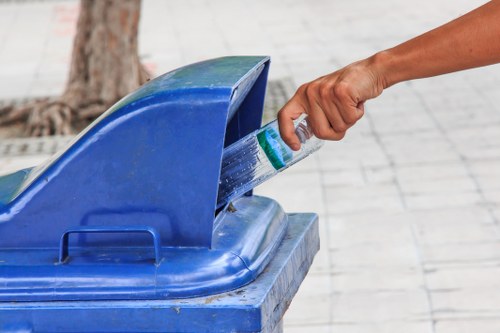 A personâ€™s hand is shown placing a plastic bottle into the open top of a large blue recycling bin, which is situated on a paved sidewalk in an outdoor urban environment. The bin appears to be made of sturdy plastic with a slightly textured finish and is positioned near the base of a tree, with visible roots and part of the trunk in the background. The surrounding area includes a paved surface, likely made of concrete or stone tiles, with some dirt and debris near the tree. The scene is lit with natural daylight, indicating an outdoor setting, and the action suggests waste disposal or recycling activity, relevant to rubbish removal services provided by Office Clearance Deptford in the vicinity of Deptford. The focus on the recycling bin and the act of depositing waste emphasizes environmentally conscious waste management and clearance efforts in the local area.