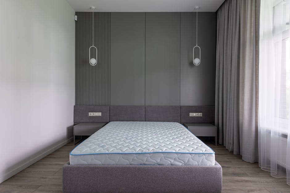 A minimally furnished bedroom featuring white walls and a wooden floor with a warm, natural finish. The room is illuminated by natural light coming through two large windows; the left window is a glas
