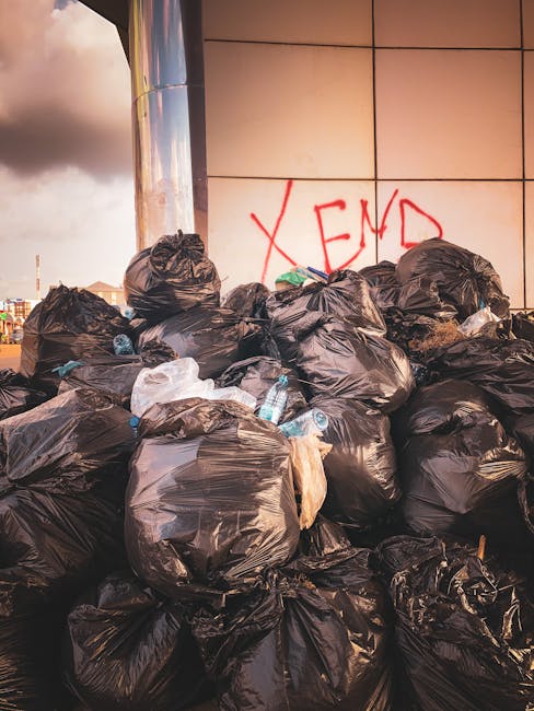 A large outdoor waste collection point situated on a paved urban street, featuring multiple overflowing bins and numerous loose rubbish scattered around. The central container is a grey mixed waste bi